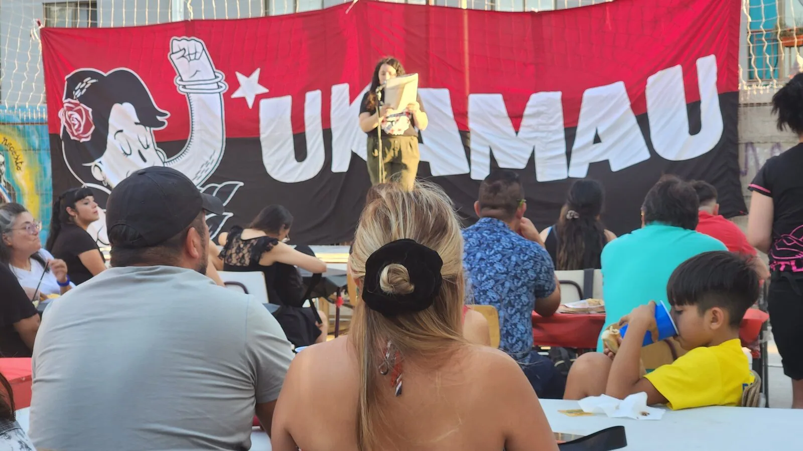 Asamblea del Barrio Maestranza Ukamau: vecinas y vecinos reunidos bajo el lienzo del movimiento mientras una compañera interviene en el micrófono