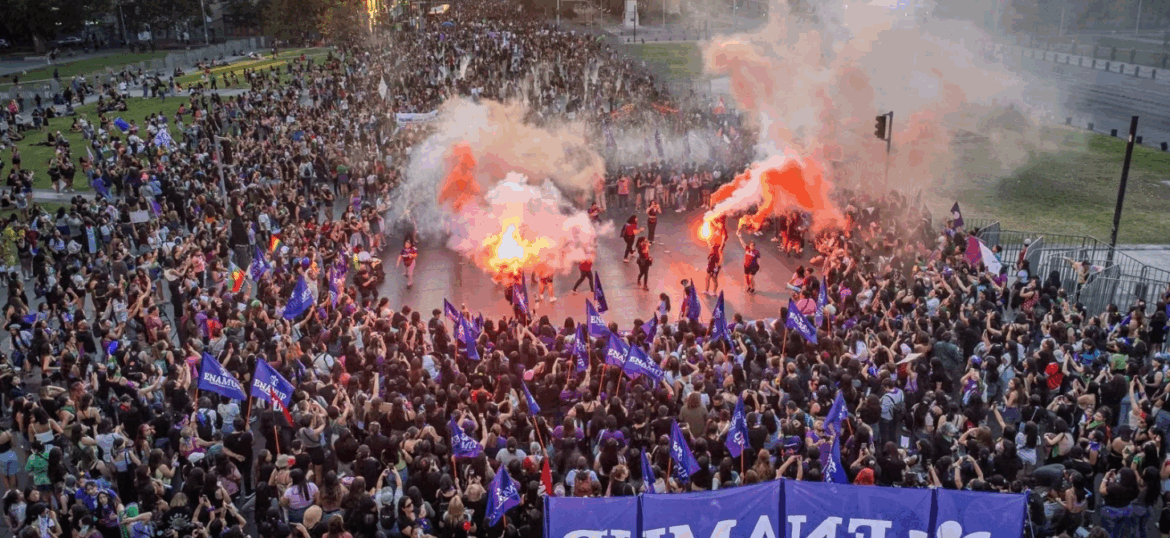 Multitudinaria marcha de mujeres de Ukamau con banderas moradas y bengalas naranjas, manifestándose por los derechos sociales y contra la violencia hacia la mujer.